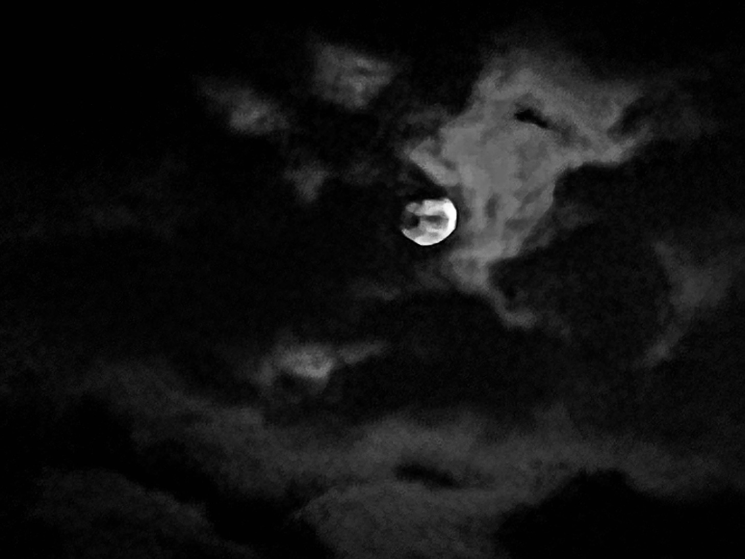 A moon shining brightly in the dark night sky, partly obscured by wispy clouds stretching across it and the night sky.