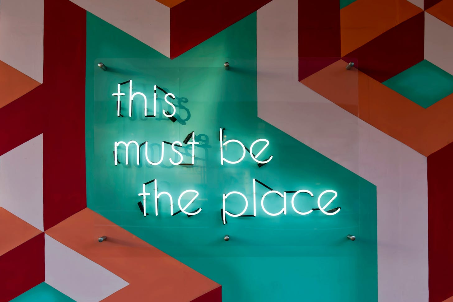 Una parete decorata con geometrie bianche, rosse, verdi e arancioni. Al centro un'insegna al neon dice "this must be the place".