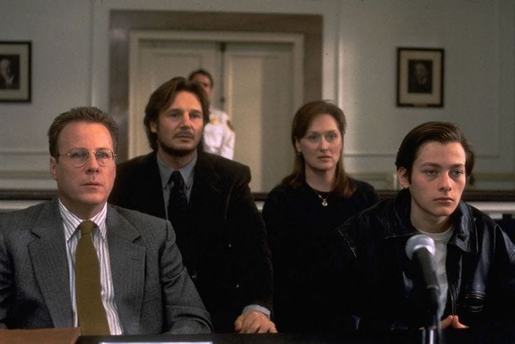 This may contain: three people sitting at a table with microphones in front of them and one person standing behind the man