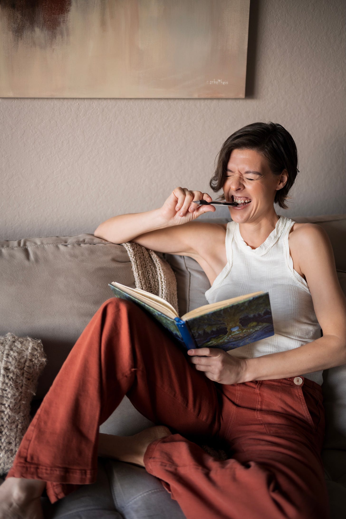 Woman with short brown hair, wearing a cream colored tank top and orange pants, sits on a couch holding a journal and biting the end of a pen.