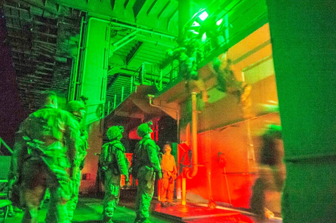A blurry shot of two Marines in tactical gear climbing ropes attached to a balcony as fellow Marines watch illuminated by overhead lights.
