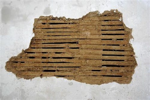 Photo of a large ragged hole in  plastered wall. Where the plaster has been removed you can see the wooden laths behind it. 