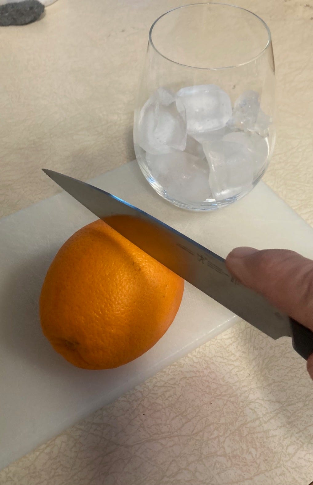 An orange on the cutting board is about to be sliced. A tumbler of ice waits for the drink.