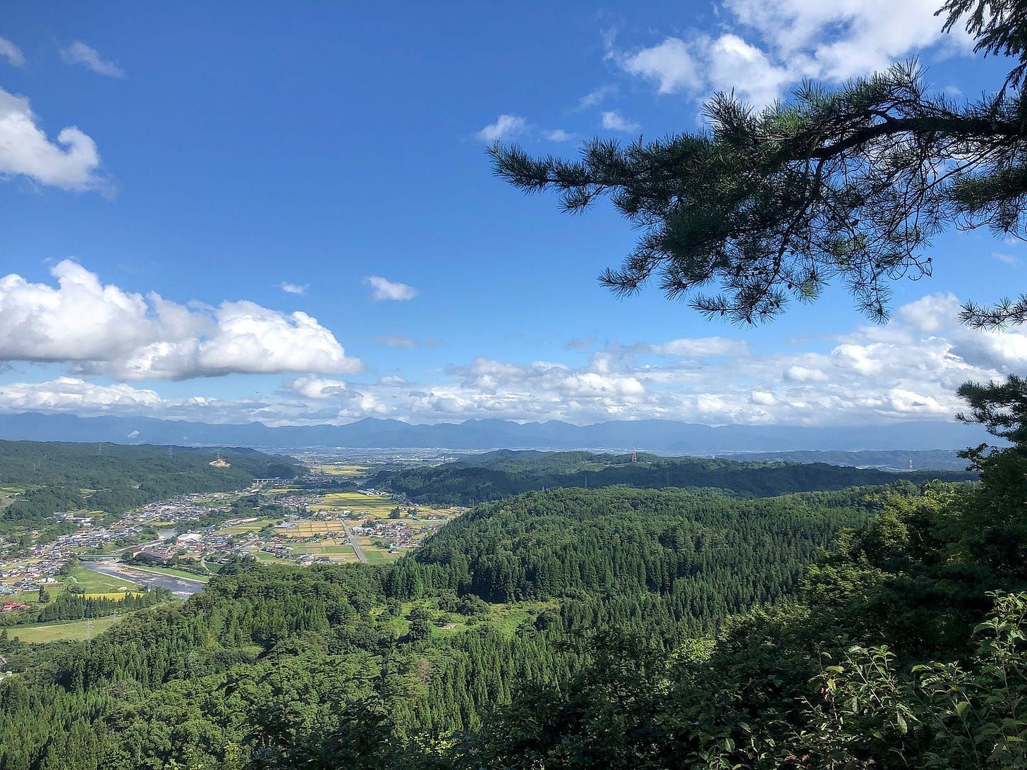 Looking out from the summit of Tengu-yama in Nishikawa, Yamagata Prefecture towards a bright blue sky with clouds, mountains in the distance, forests and rural towns in the foreground.