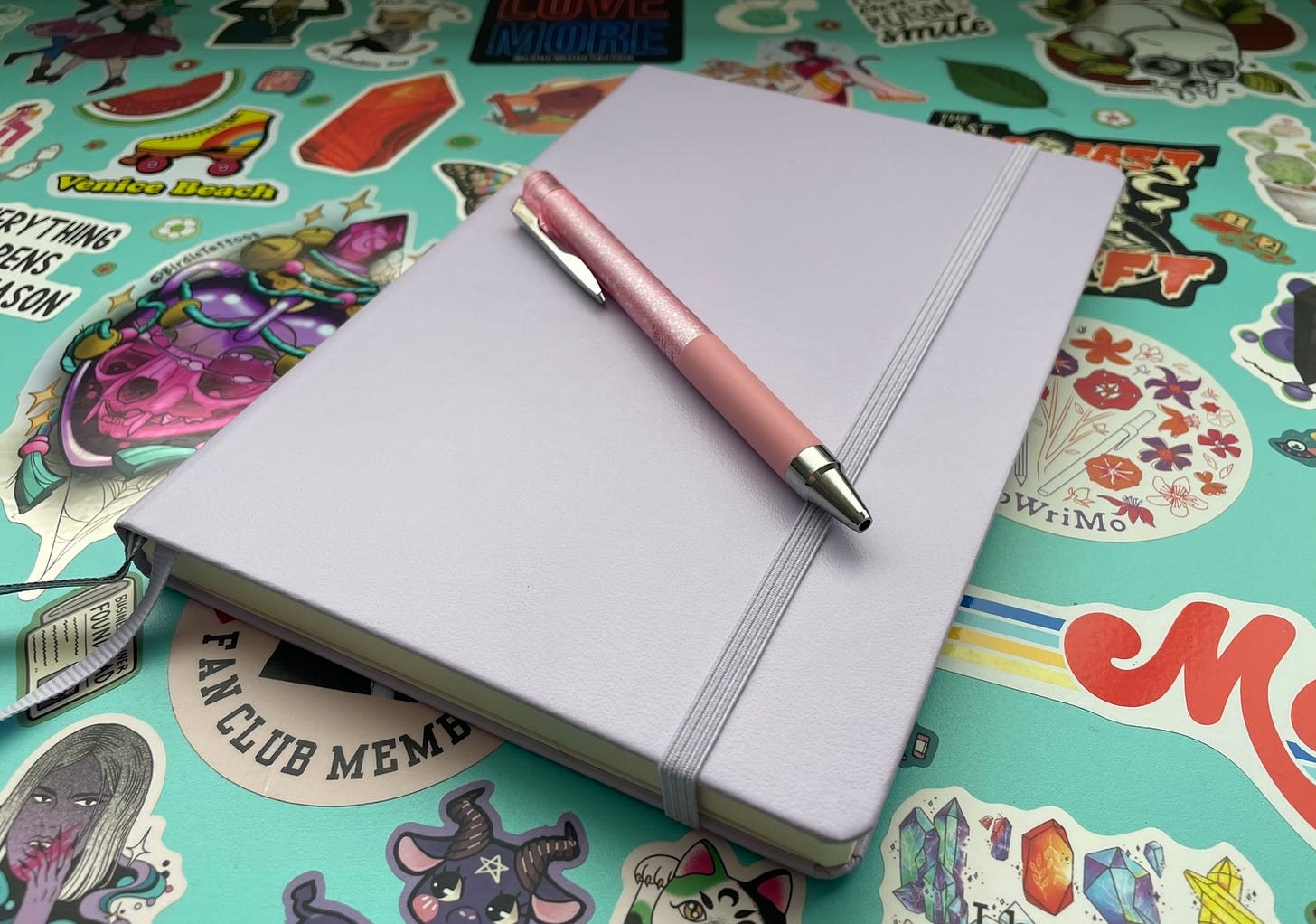 Image is of a pink pen sitting on top of a pale lavender coloured notebook. Both are sitting on a turquoise coloured desk that's covered in stickers.