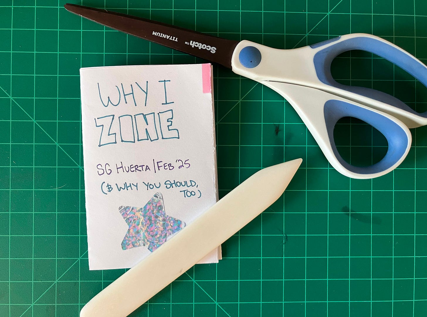 A copy of SG's zine sits on a green cutting mat. A bone folder rests on top of the zine and a pair of scissors is next to it. The zine is a small, folded white sheet of paper.