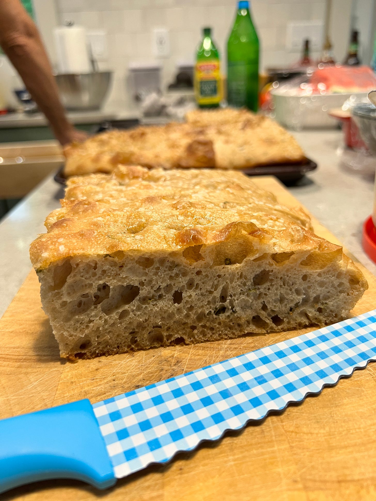 Two freshly baked loaves of focaccia bread are sitting on a counter with various cooking implements and olive oil in the background.