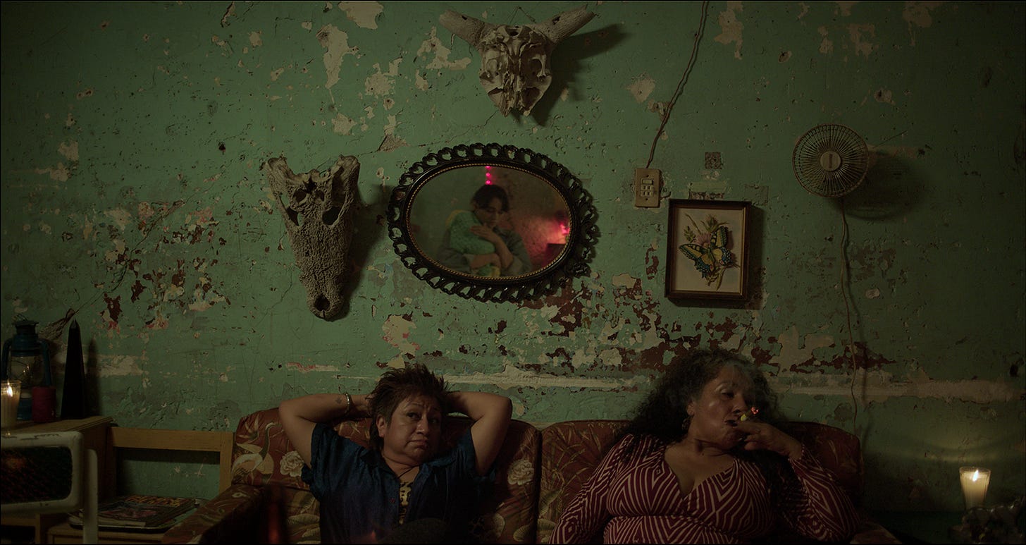 In a cement-walled, peeling-paint room, two women sit on a couch below a mirror, two skulls, and a framed artwork. The woman on the right side of the couch is looking down as she takes a drag on a cigar. The woman on the left rests her arms up above her head while she looks up and to the left, ostensibly at the woman reflected in the mirror who is standing holding a tiny baby wrapped in a green-and-yellow knit blanket.