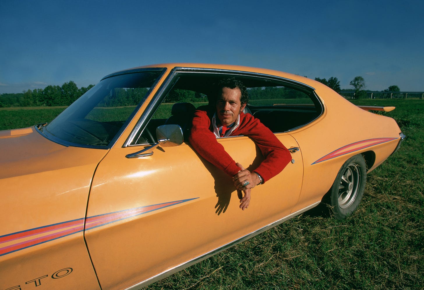 Photo of a man sitting in a GTO car parked in a grassy field.