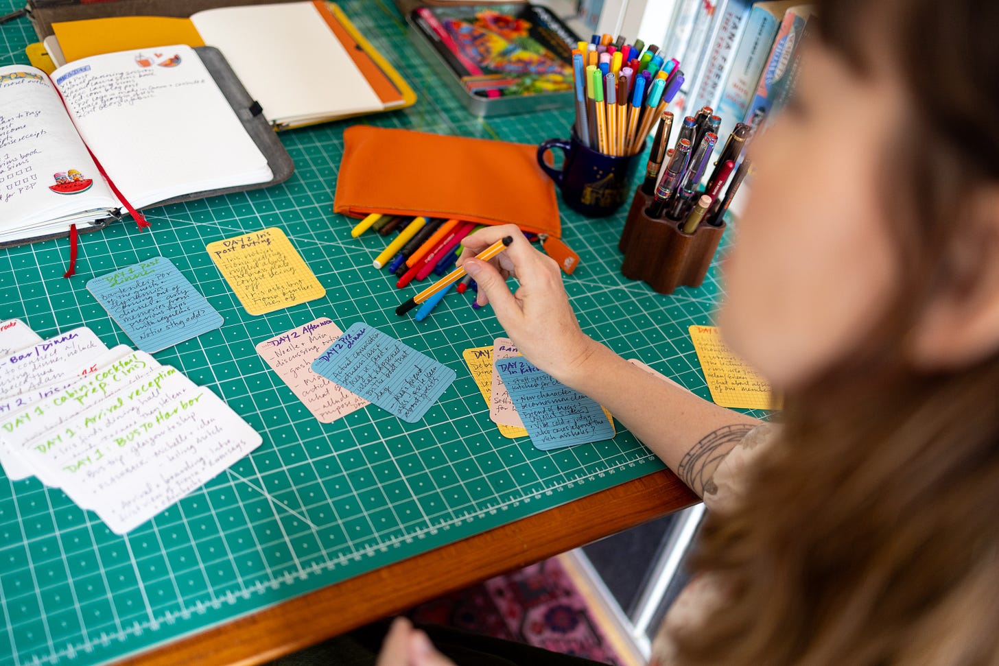 A woman with a tattoo on her forearm looks at a desk with a green blotting pad covered with colorful notecards and open notebooks with writing inside. She's got a bag of colored pens open. 