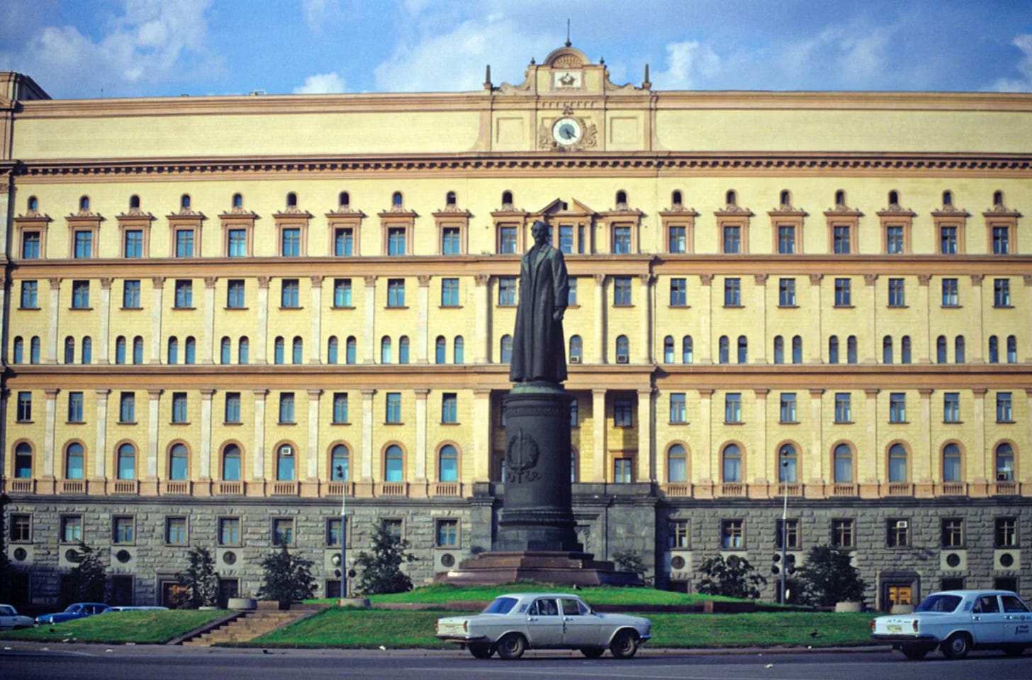 A stolid-looking yellow building, et on a foundation of stone, looms over a small grassy traffic circle, below. A black statue of the founder of the KGB glares at passing cars. 