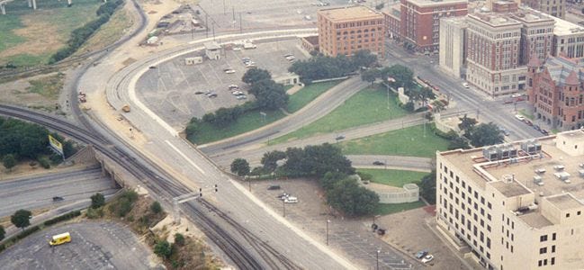 Photo of Dealey Plaze. A large central road runs more or less veritcally. To the left is more roadway and an open field, to the right a number of parkings lots and boxy structures