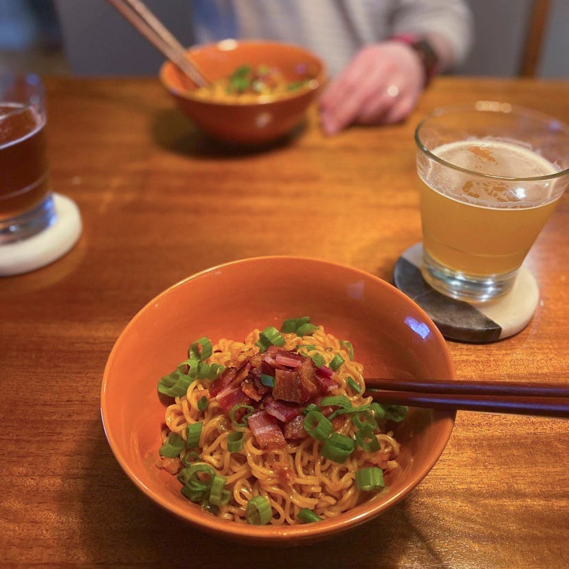 Two orange bowls with chopsticks sticking out of them, across from each other on the table. Each bowl has ramen noodles in an orange sauce, and piled on top are pieces of chopped bacon and sliced green onion. Two glasses of beer sit on coasters near the bowls.