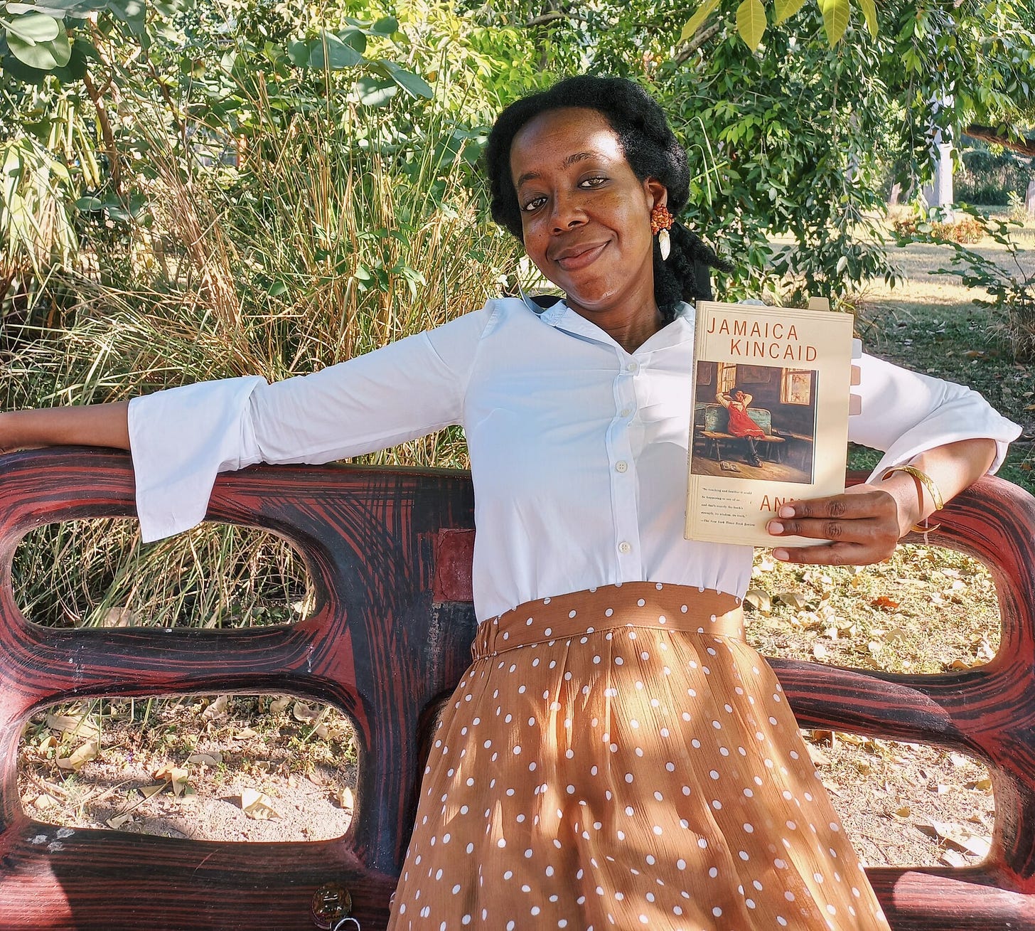 A black woman with natural hair in a loose ponytail, wearing a light brown skirt with white polka dots and a long sleeve white shirt is sitting on a red bench under a tree surrounded by greenery. She's holding a paperback copy of Annie John as she leans back against the bench rest with a slight smile.