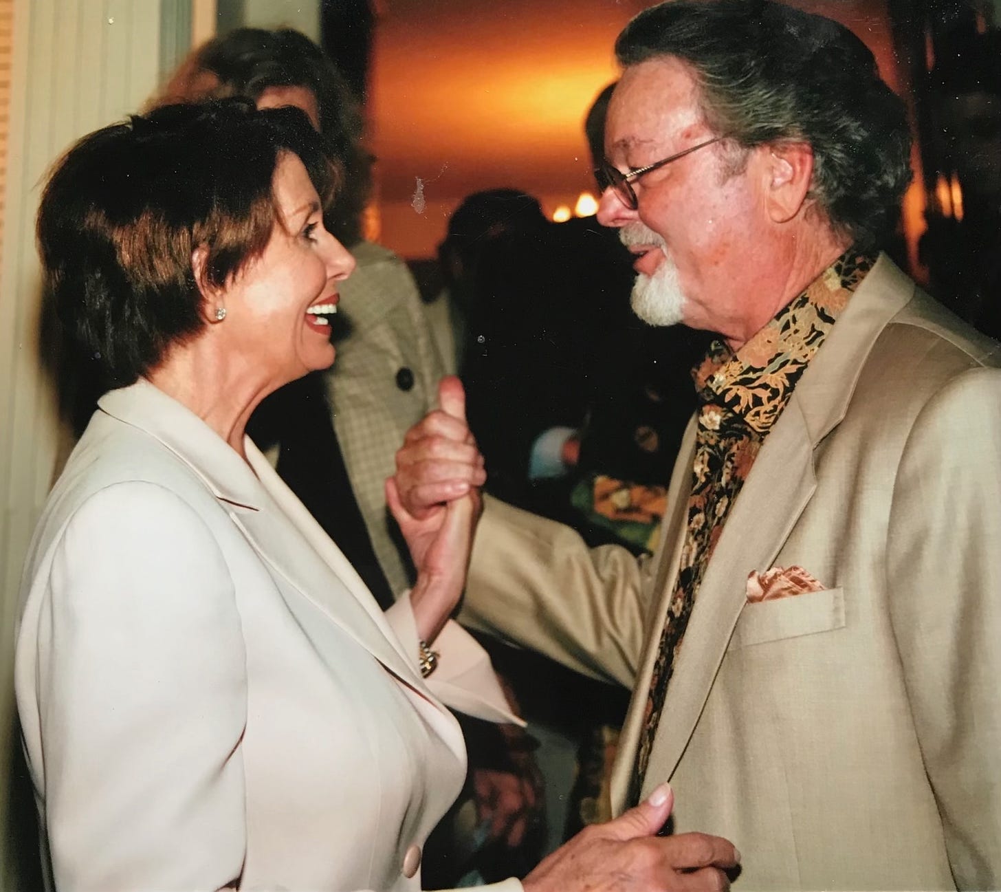 Nancy Pelosi and Russ Tamblyn clasp hands while they speak to each other at an event.