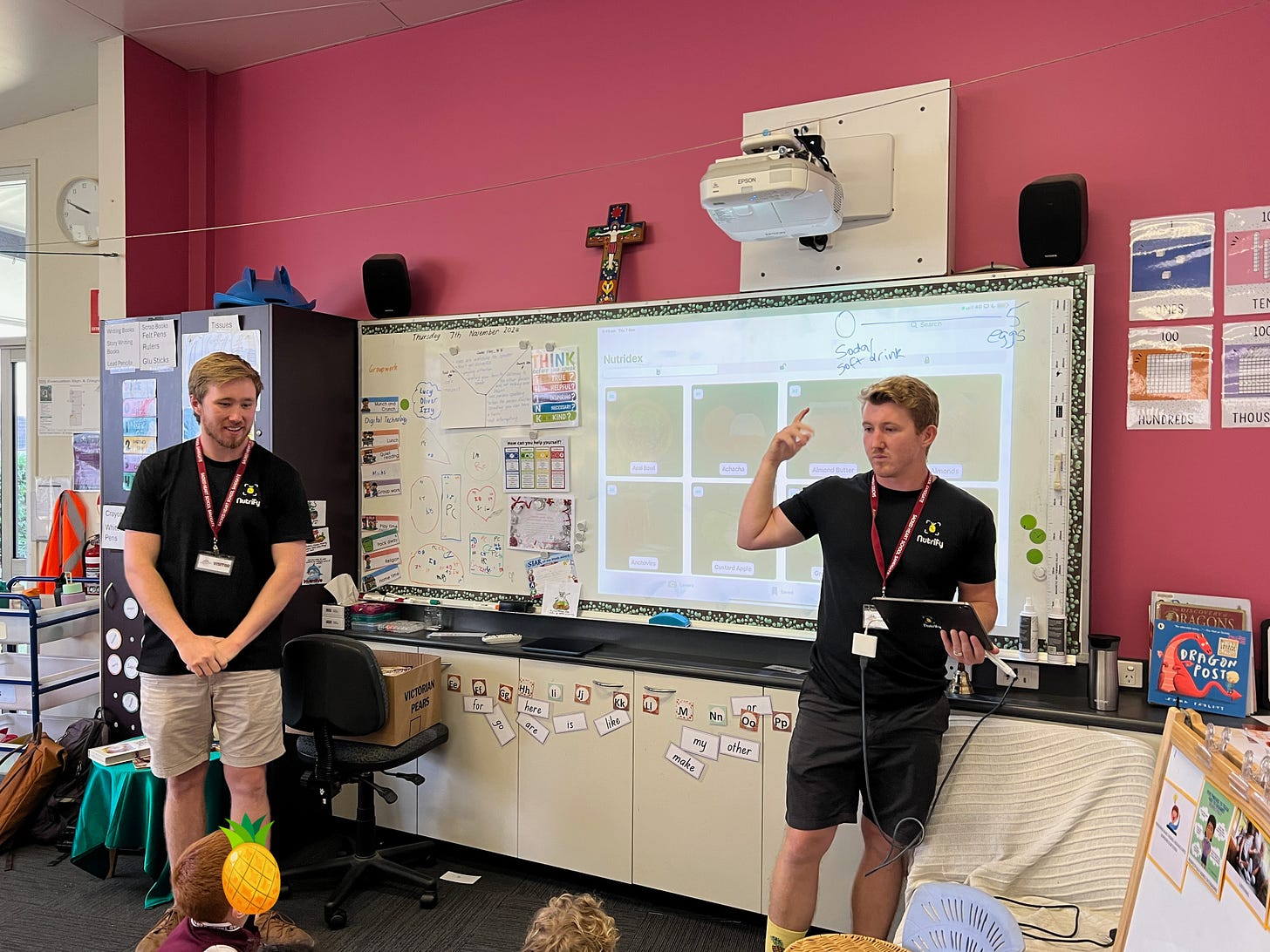 Two men wearing Nutrify shirts and red lanyards present to a classroom of young students. One holds a tablet, gesturing while explaining, and the other stands smiling. The classroom has a pink accent wall, a whiteboard with Nutrify app content displayed, and various educational materials and posters surrounding the board.