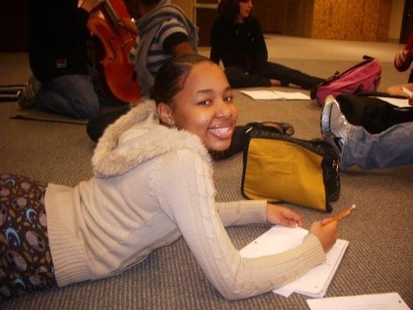 A black high school girl laying on the carpeted floor writing in a notebook and smiling at a camera with  people chilling and holding a base in the back.