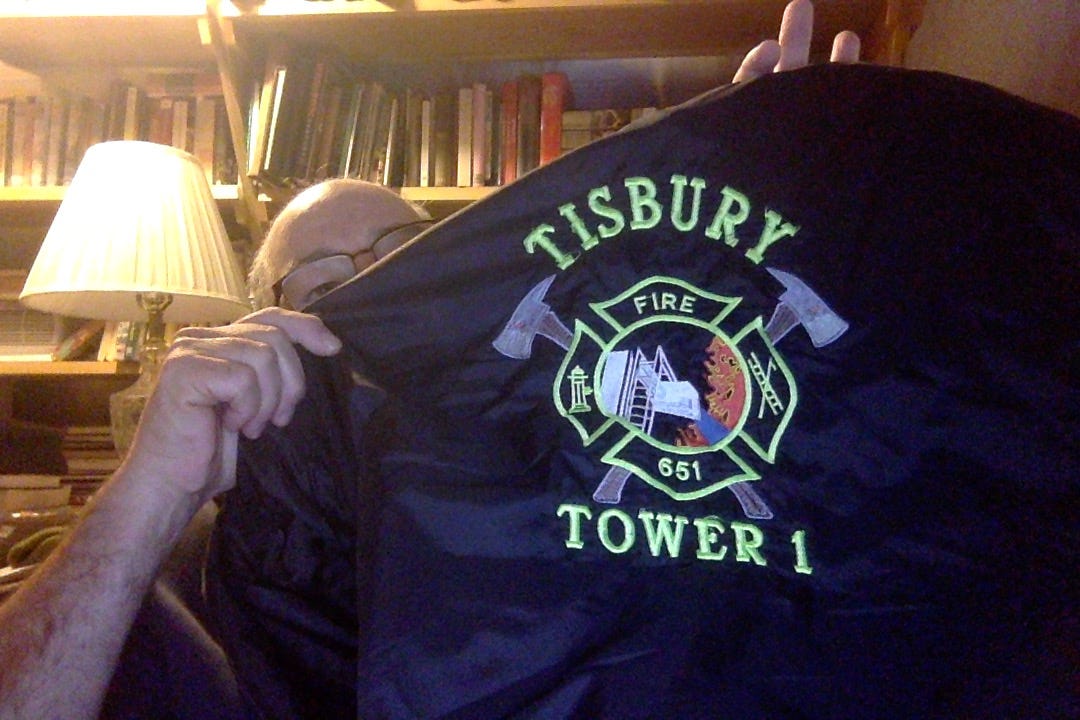 Photo of me holding my machine-embroidered windbreaker.  Above and below an image of a tower water cannon shooting blue water into scary flames, a maltese firefighter cross, with crossed axes, an image of a hydrant, a pike pole and a ladder and a hydrant and the words "fire" and "651"
