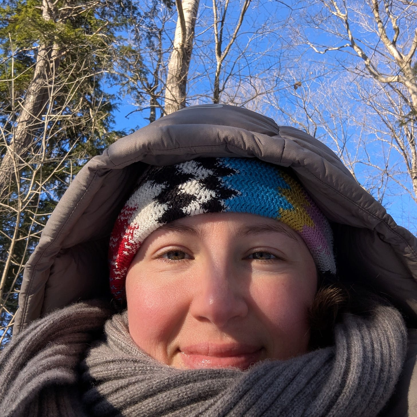 Aleksa stares at the camera, bundled up in a taupe coat, grey scarf and multicoloured hat, smiling with red cheeks, with trees and the blue sky behind them.