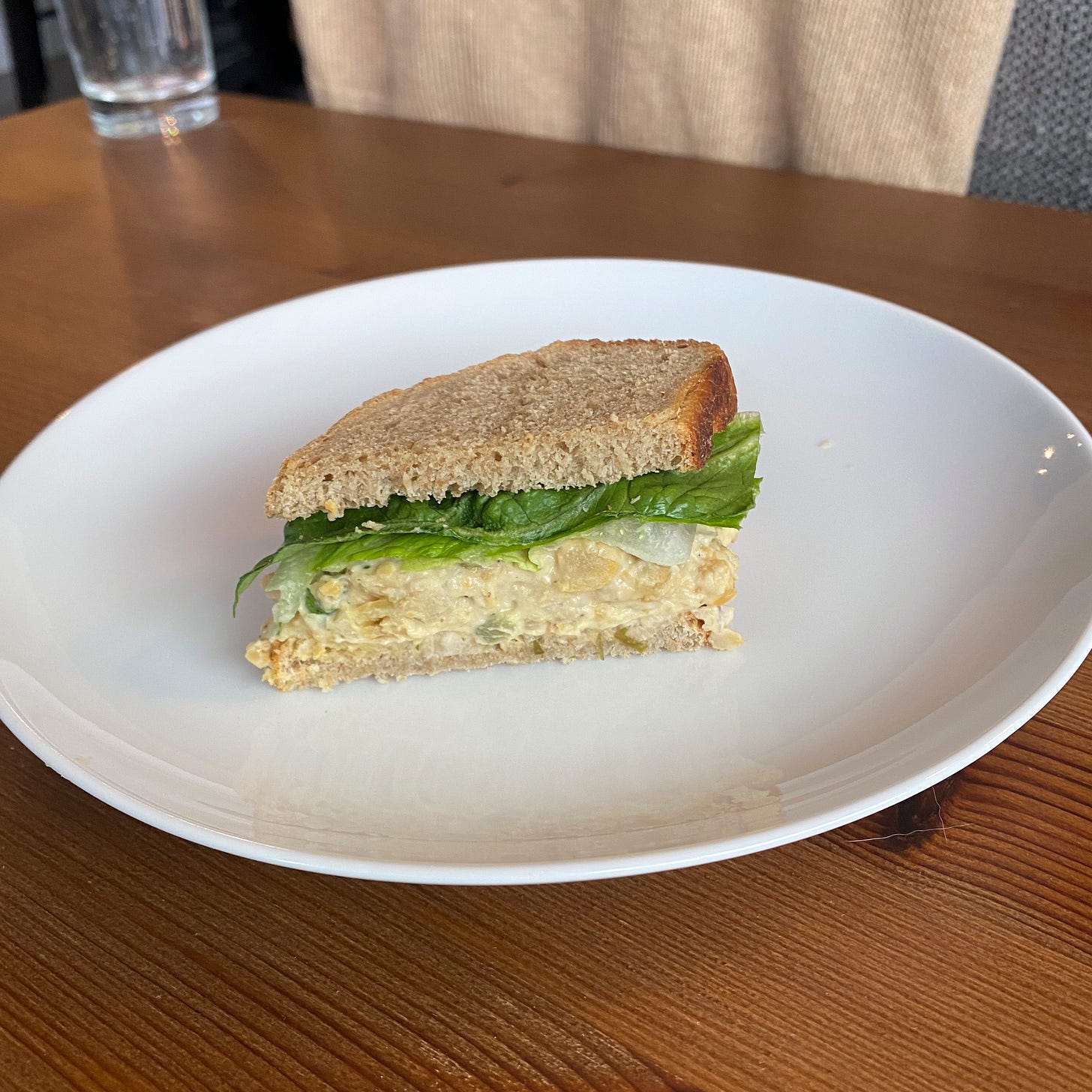On a white plate on top of a wooden table, half of a chickpea 'tuna' salad sandwich with lettuce. The bread is light brown with a dark crust.