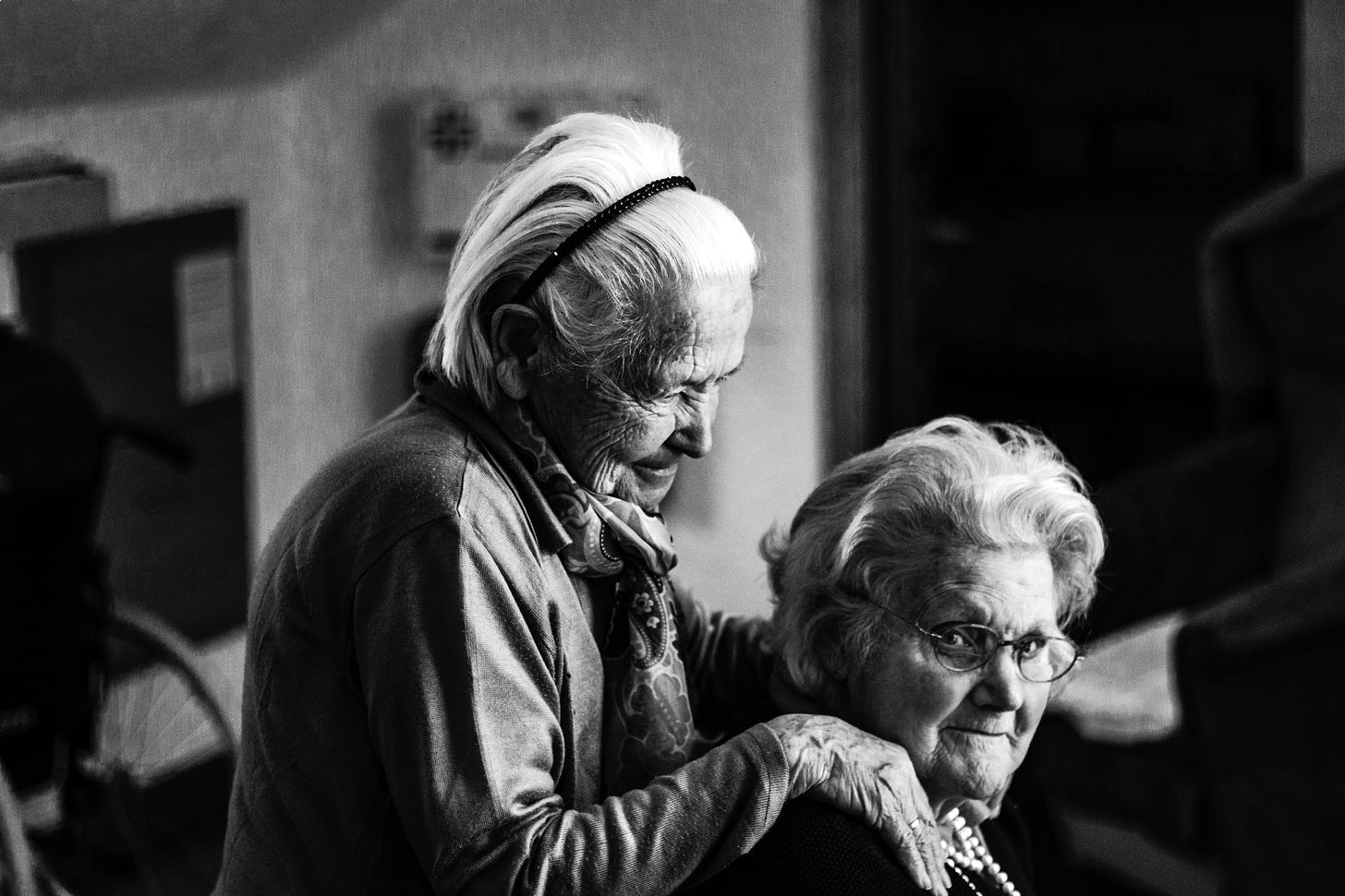 Foto de duas mulheres idosas, uma delas segurando no ombro da outra, numa foto preta e branco. 
