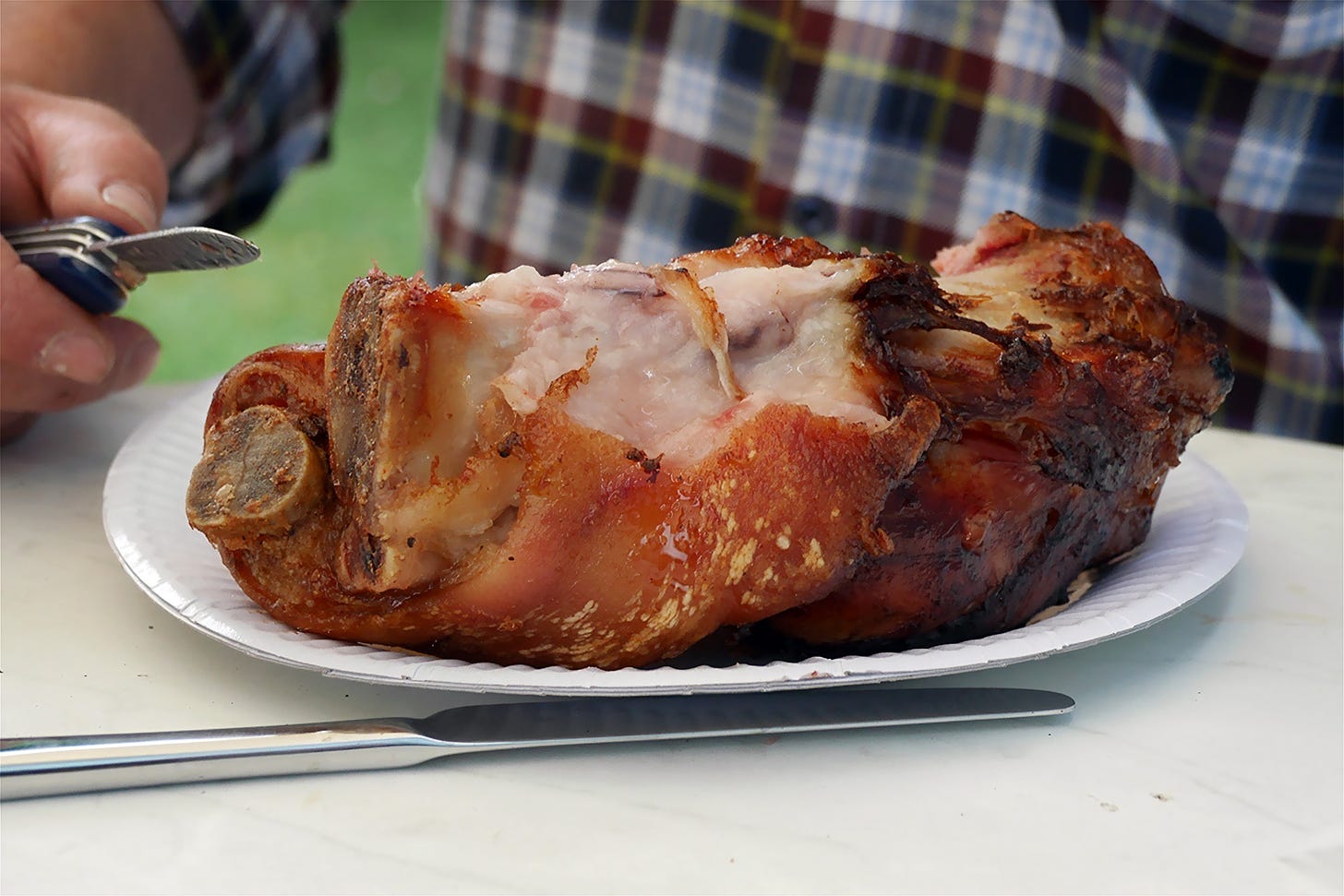 A close-up image of a large, greasy German pig knuckle served on a white paper plate. The pig knuckle has a crispy, golden-brown exterior and tender, juicy interior, with some visible fat and meat. In the background, a person wearing a plaid shirt is holding a knife, ready to cut into the meat. A stainless steel fork and knife are placed on the table next to the plate. The setting appears to be an outdoor dining area, possibly a beer garden, with a blurred green background. Public domain.