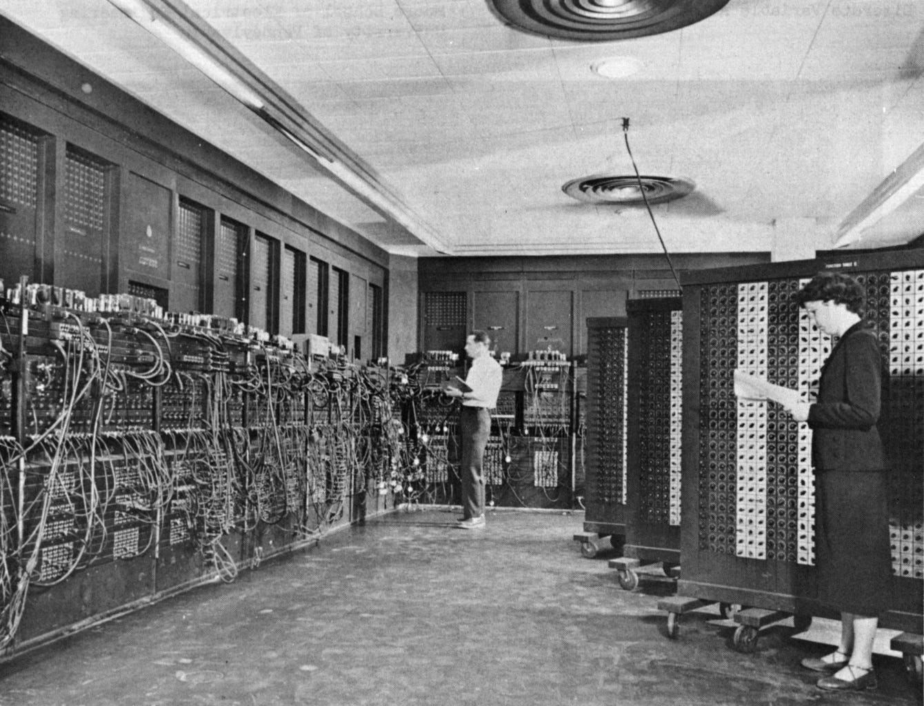 Black and white image of two programmers standing in front of an array of vacuum tubes and wires.