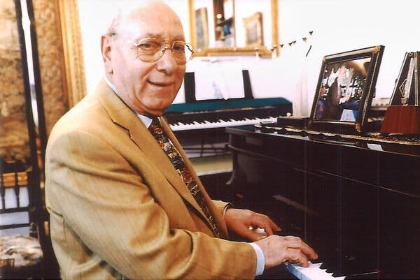 A picture of a bald older man in aviator-style eyeglasses and a tan suit jacket sits at an upright piano, his hands on the keys, in what appears to be a private home.