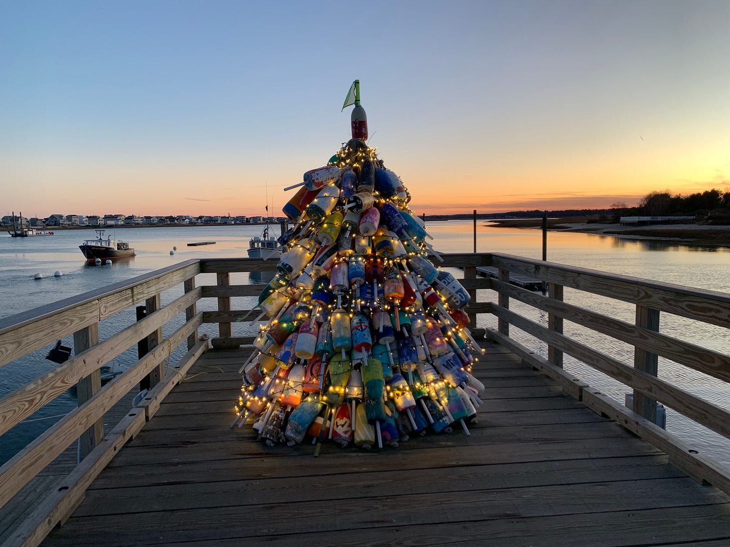 A christmas tree made of buoys is sitting on a pier at dusk. It is lit with white lights and the buoys are all colors. The sky is blue, peach, orange and red. The water is rippling slightly.