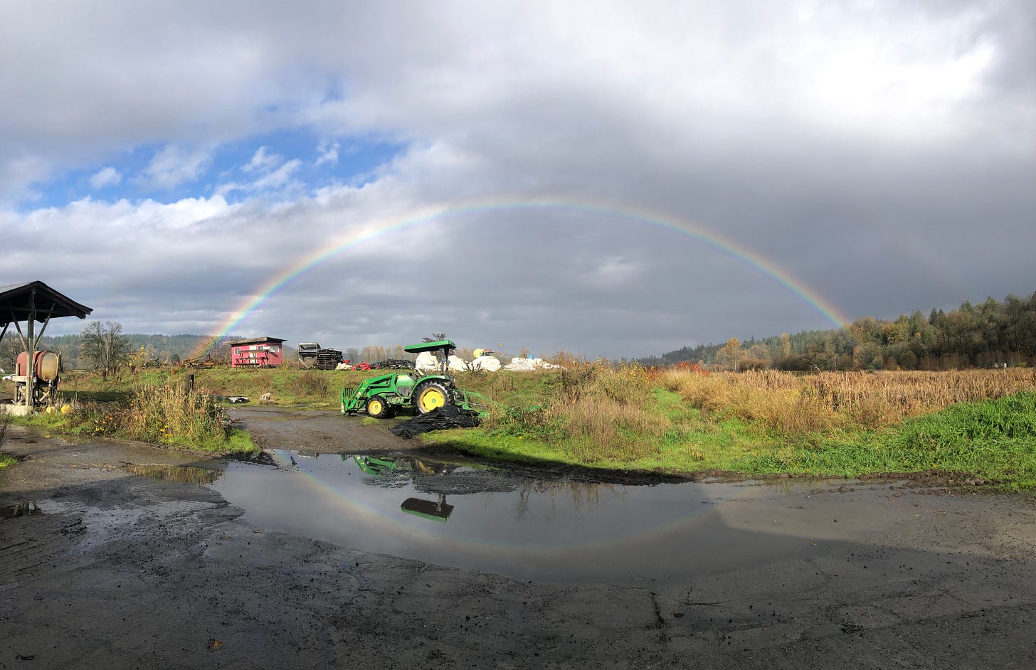 a full rainbow, stretched end-to-end, over our farm, reflected in a puddle below, with a bright green tractor in the foreground