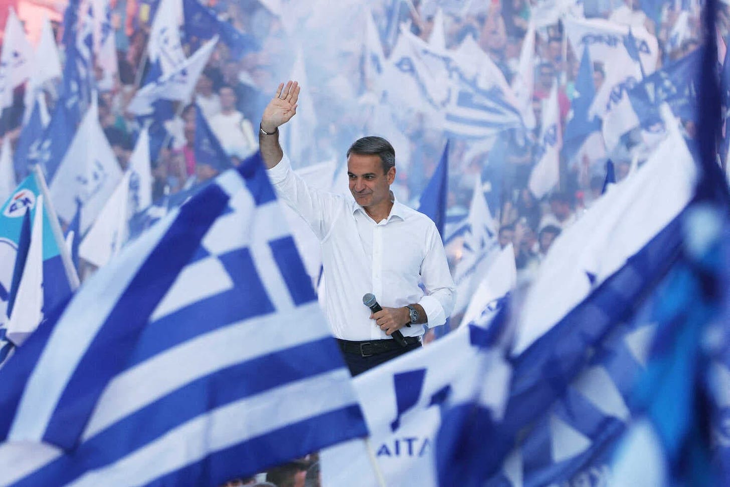 A man in a white dress shirt waves to a crowd while holding a microphone amid a sea of blue and white flags.