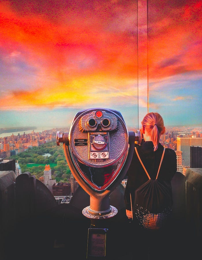 A set of binoculars overlooking a city at sunset, with a woman looking over it to the side.