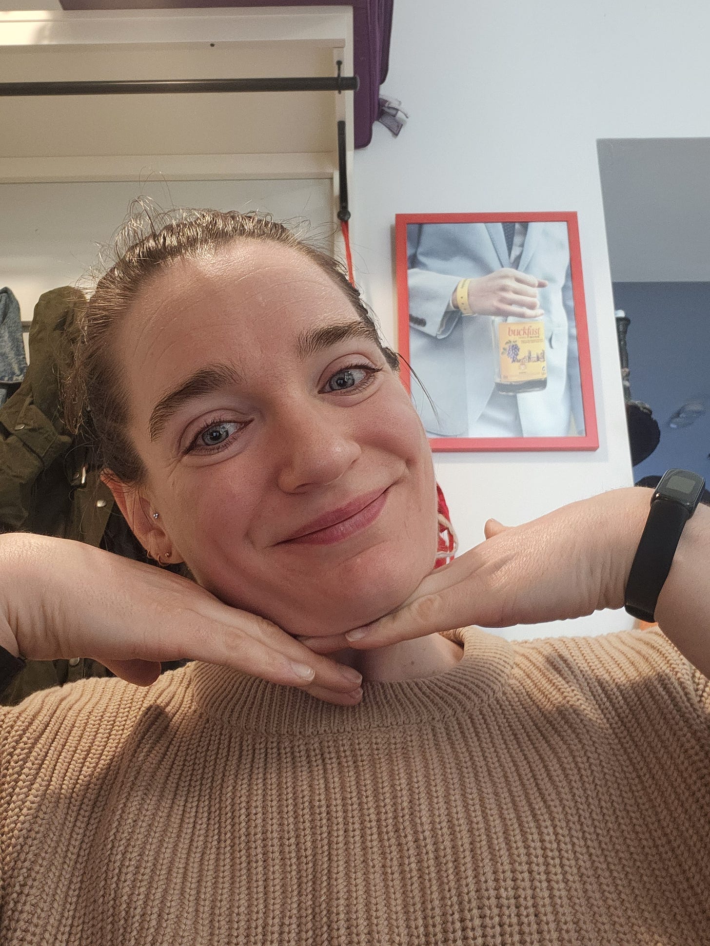 Smiling like an angel, and with two hands posed under my face like a child star, this is a selfie. My hair is pulled back into a bun, and behind me a framed poster of a Buckfast bottle looks on. 