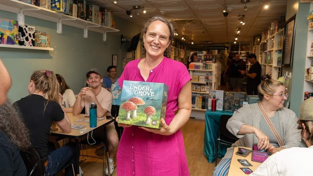 Elizabeth Hargrave, holding a copy of Undergrove, standing in front of a room full of people playing Undergrove