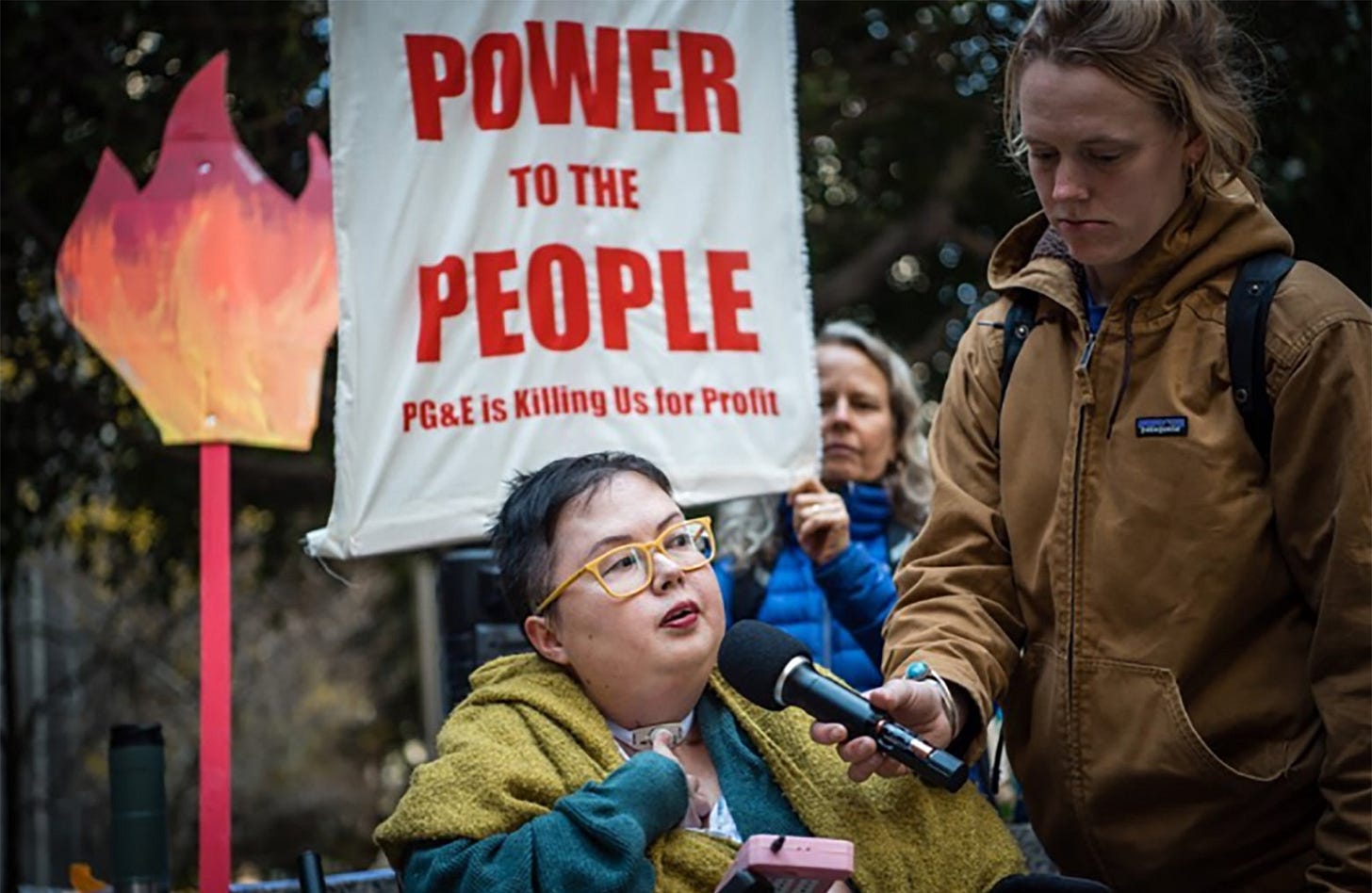 Stacey Milbern, a Korean. white powerchair user wearing glasses, stands with a friend holding a mic up to her face in front of a banner that reads POWER TO THE PEOPLE/ PG&E IS KILLING US FOR PROFIT.