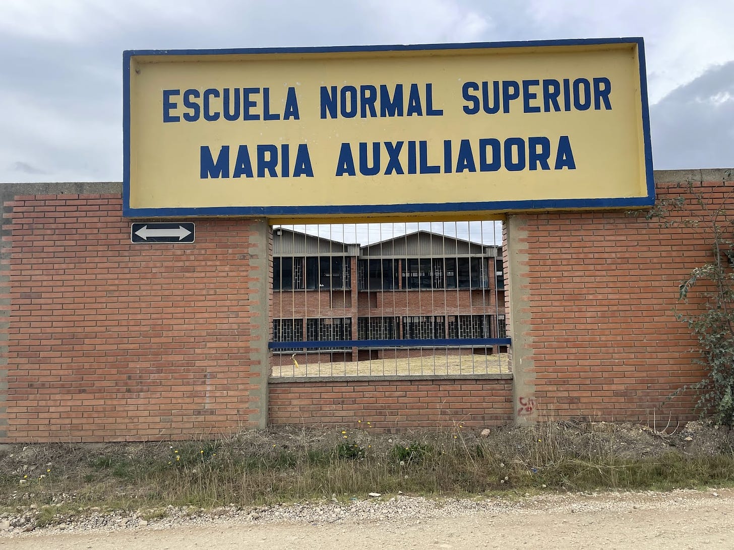 a large school sign that reads Escuela Normal Superior Maria Auxiliadora placed on top of a tall brick fence, with a portion of bars in the center, revealing a school building in the background