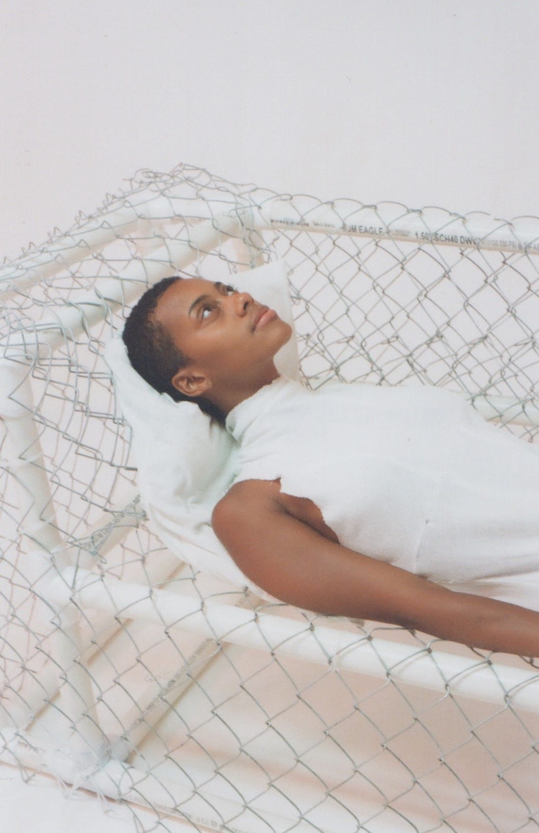 A black woman laying on a couch made of chain link fencing wearing a white bodysuit.