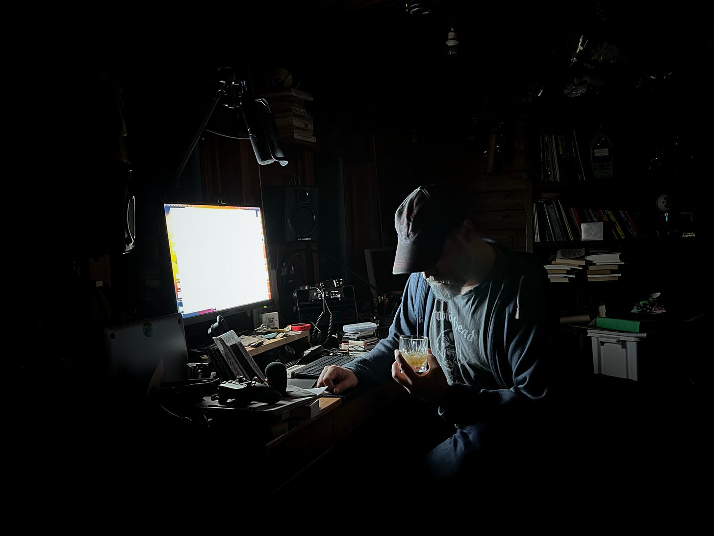 A photo of me in the shed. lit by the glow of a screen and placing down intro a glass of whistle pig whiskey.