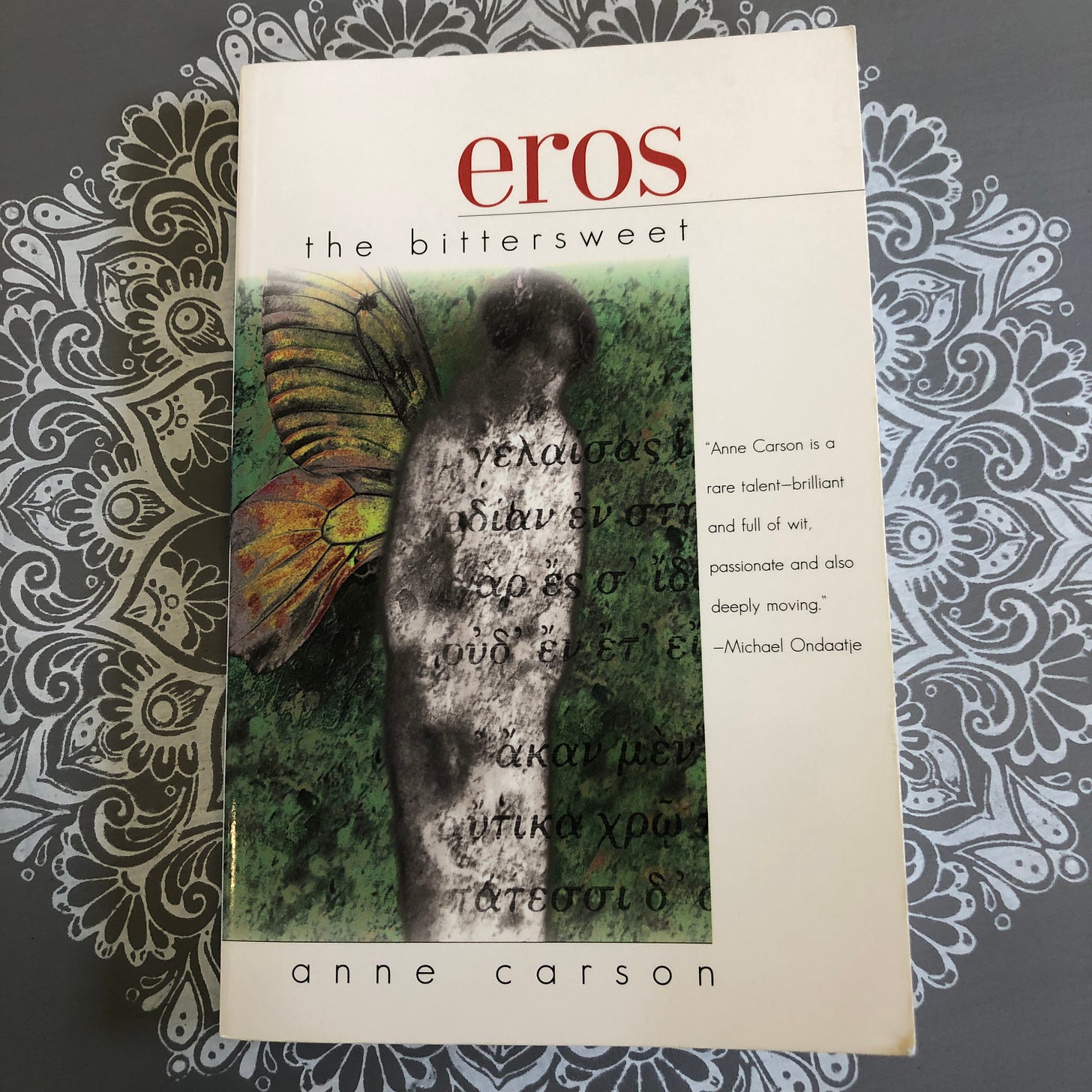 Book on table shows a cover portraying a winged creature with Greek writing superimposed and the words: “Eros the bittersweet” and “Anne Carson is a rare talent—brilliant and full of wit, passionate and also deeply moving.” —Michael Ondaatje.” And “Anne Carson.”