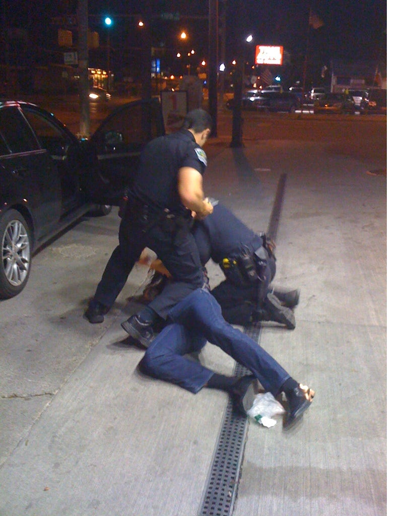 A picture from the night I was first arrested. I am lying partially on my back and partially on my side, as two cops hover over me, one with his hands on me, the other with a knee on me. It is dark because it was during the early morning hours of January 1, 2012.
