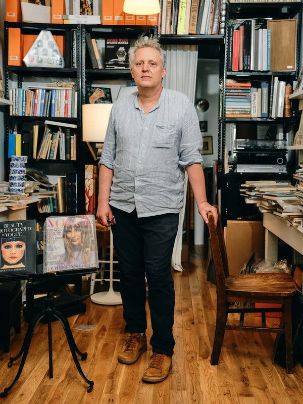 Bill Hall, wearing a rumpled blue button-down and dark pants, poses beside a chair in a room cluttered with books and magazines piled on shelves.