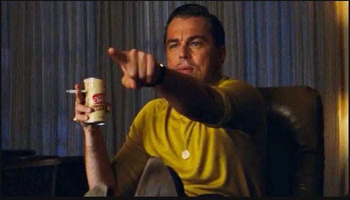 Leonardo DiCaprio holding a beer and a cigarette pointing whilte wearing a yellow shirt. 