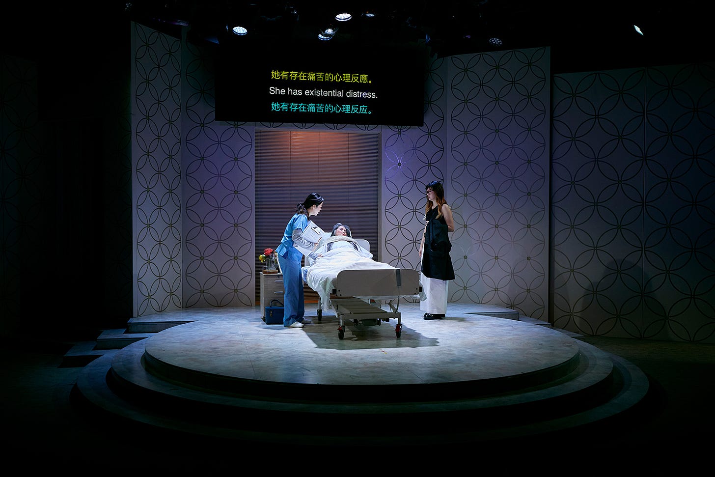 A photograph of the opening scene of the play "Miss Peony". The character Lily stands by her grandmother, who is in a hospital bed and being attended to by a nurse. Above them, there is a TV screen showing surtitles in English, Simplified and Traditional Chinese. The English reads "She has existential distress".