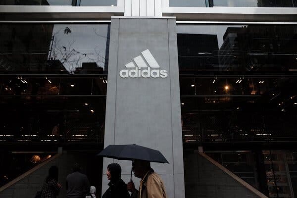 Two people under a black umbrella walk past a building with darkened glass and a column with the Adidas logo in white.