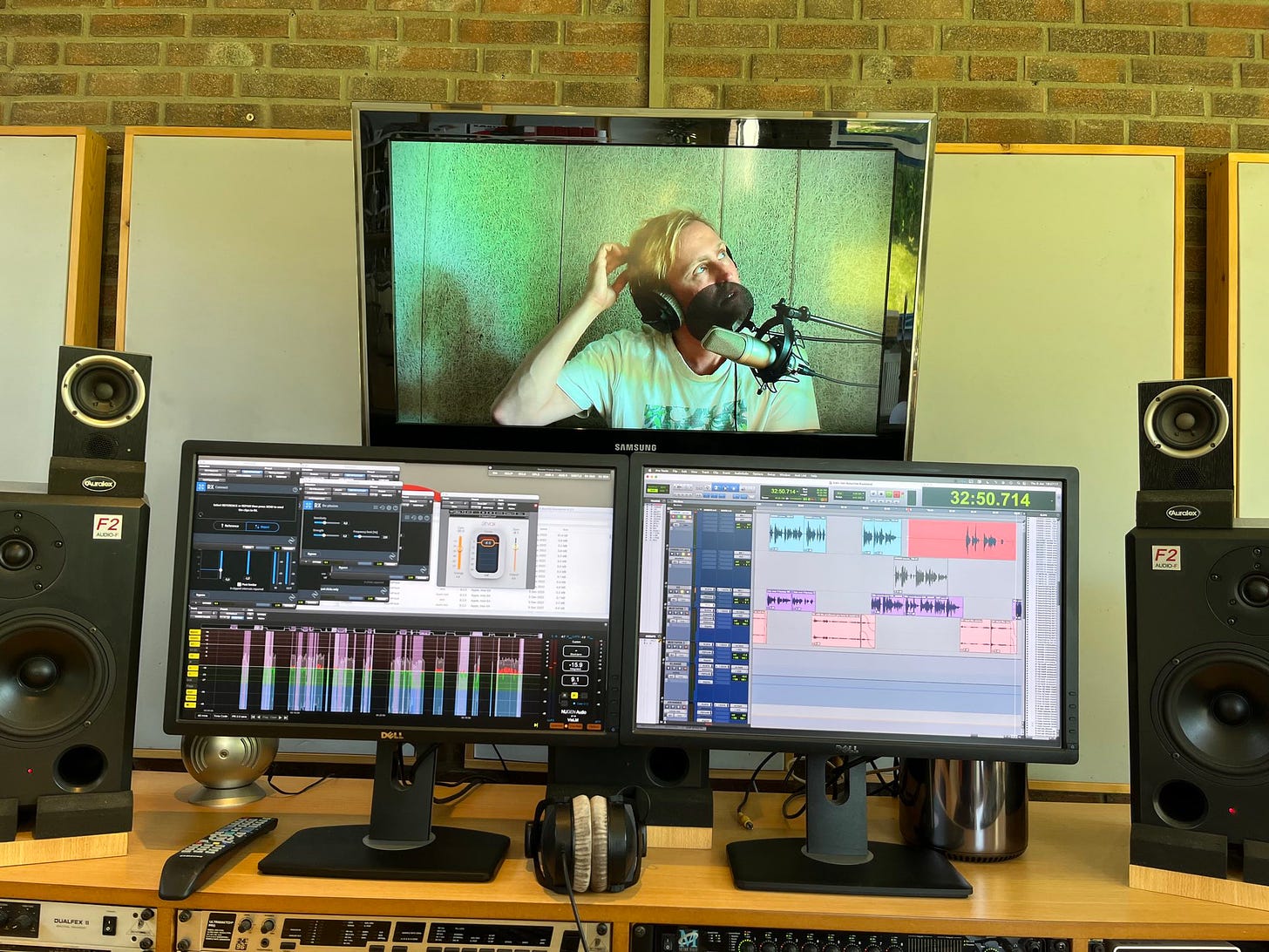 Foto van een bureau met daarop twee schermen met montage software er op, twee speakers en een grote TV erboven waarop je lieven ziet zitten in achter een microfoon met een koptelefoon op.