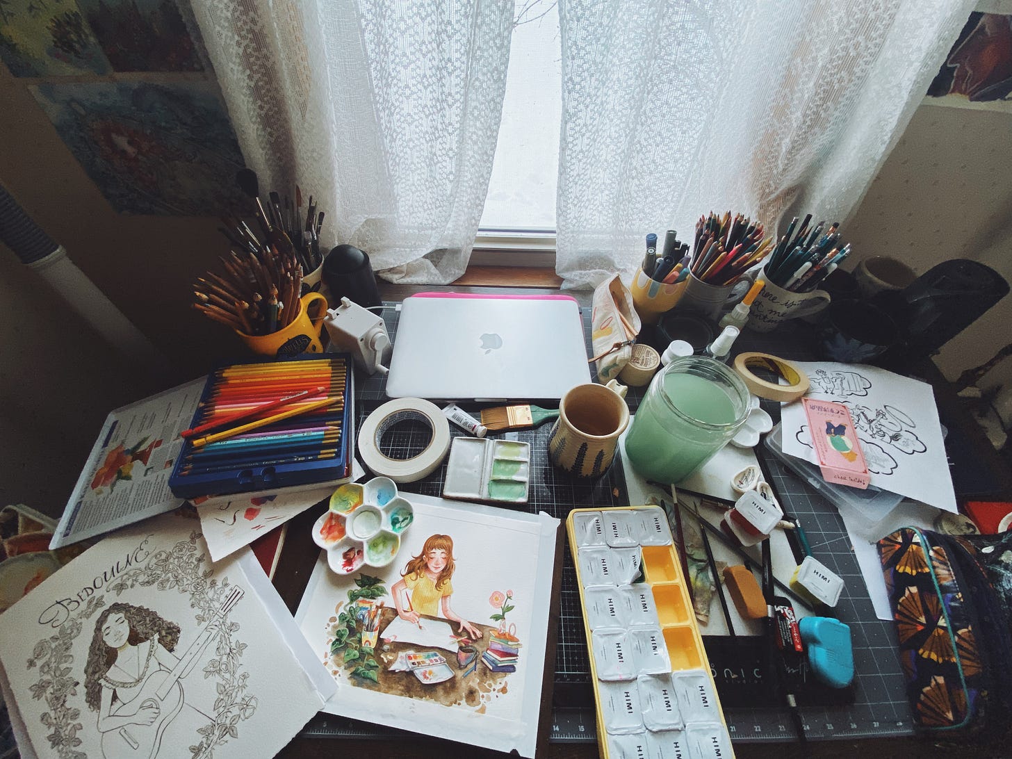 A photograph of the studio with art supplies scattered across a drawing table. Two incomplete illustrations are surrounded by the many pencils, paints, paintbrushes, and other materials.
