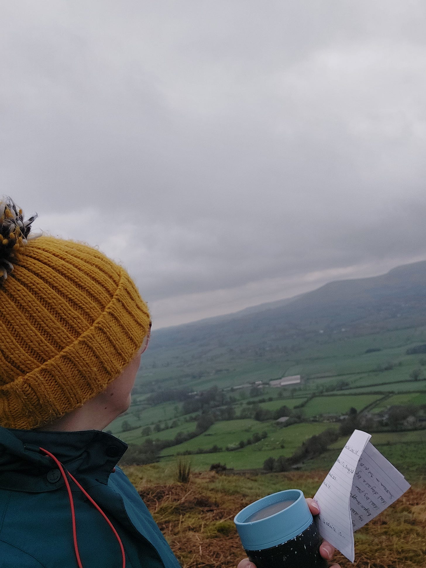 Person wearing a yellow hat gazing at out at a misty rural view of green fields. They are holding a cut and a piece of paper with journal prompts on it