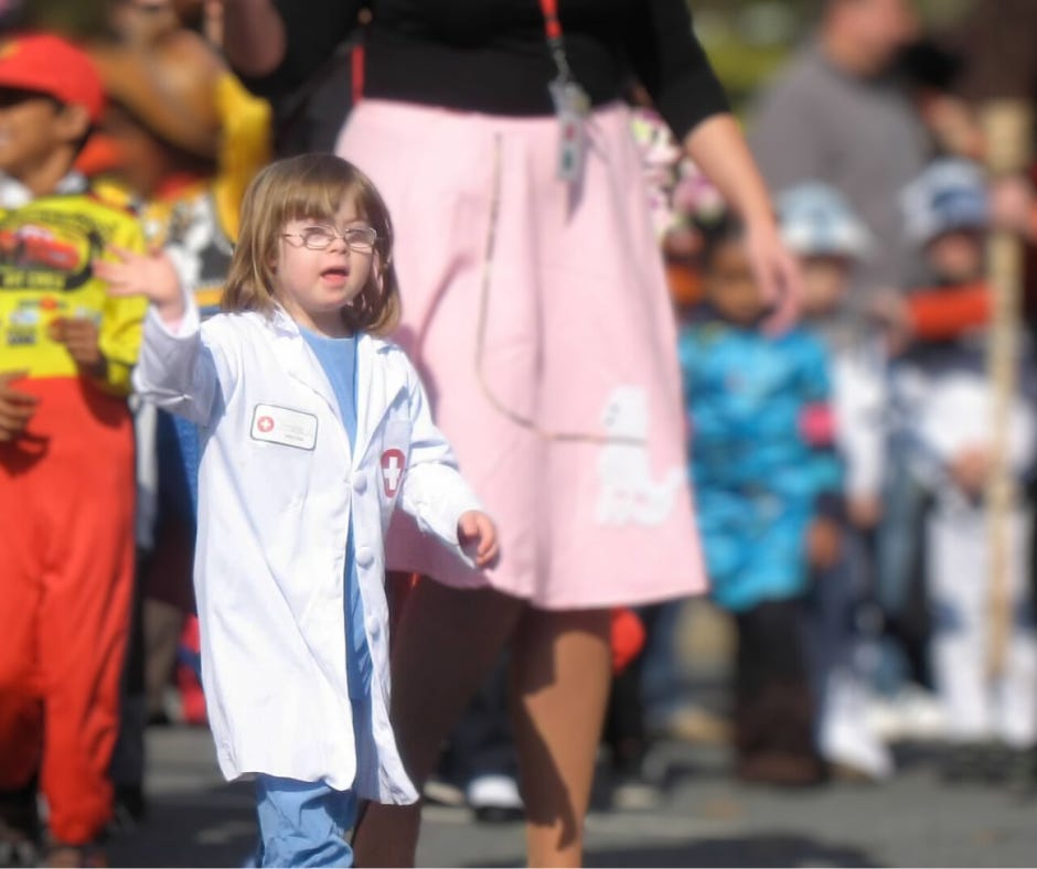 A photo from 2011 of Penny, who has Down syndrome, dressed as a doctor in a white lab coat, walking and waving during a parade. She is participating with other children, who are dressed in various colorful costumes.