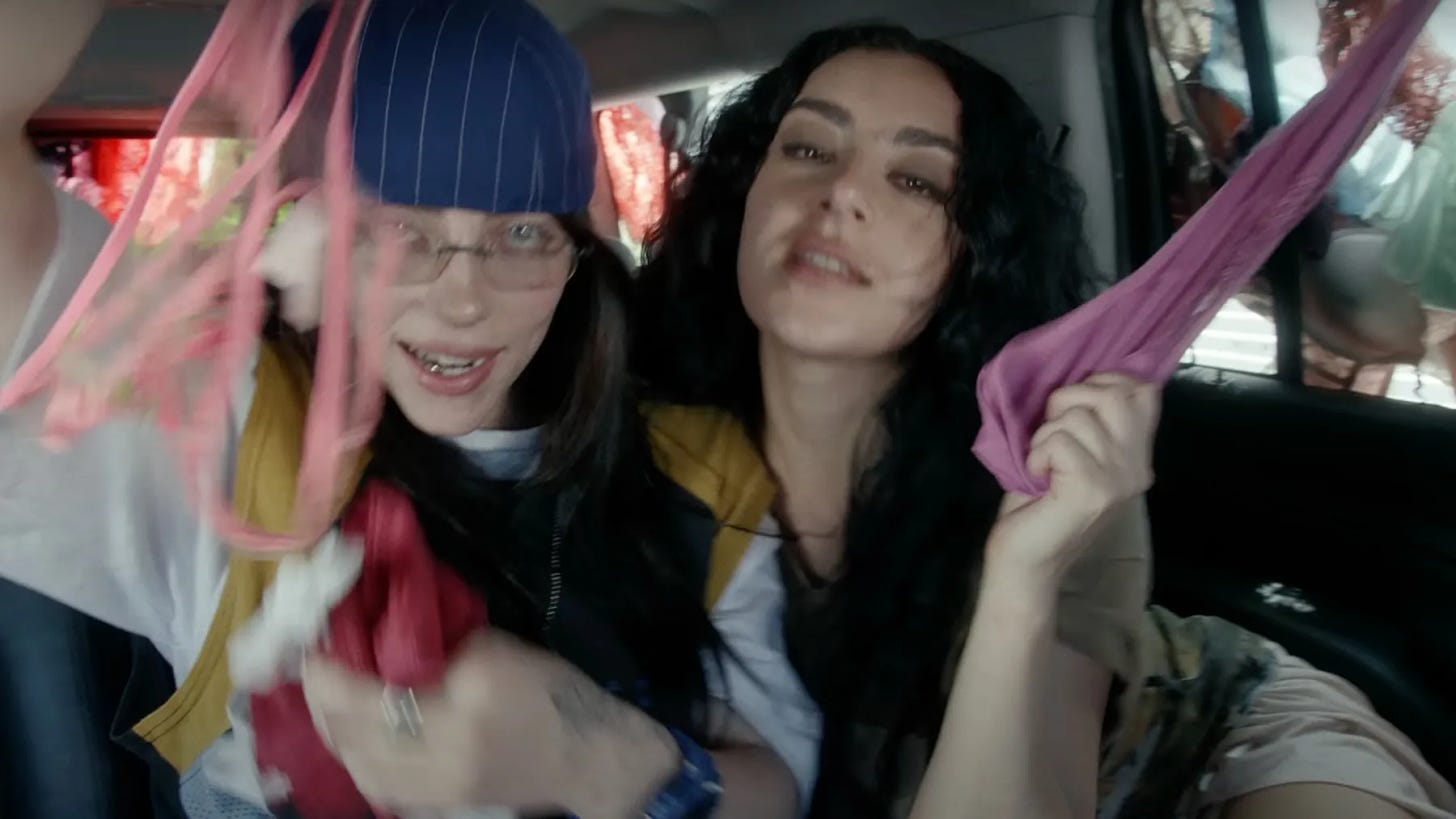 Two people pose with handfuls of lingerie in a car. The person on the left has long brown hair and wears a blue hat with white stripes and glasses. The person on the right has long curly black hair.