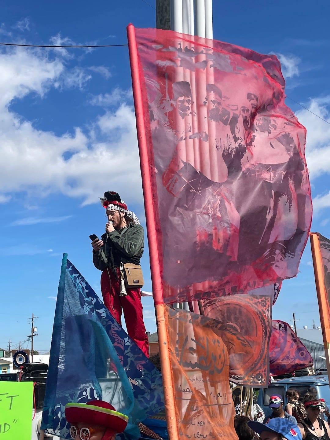 Krewe of Chickpeas speaker Marco Saah is addressing the crowd. He is standing on a box speaking into a microphone and wears a black and white keffiyeh on their head, a green jacket, and red pants. In front of them is a silk-screened flag of a traditional Palestinian band.
